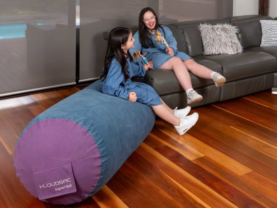Two sisters sitting next to each other in the lounge room playing with dolls, one on the couch and one sitting on the KloudSac Supersub.