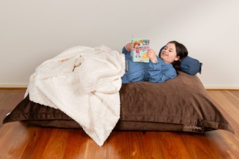 Young girl reading a book while lying on a KloudSac pillow with a polar calming blanket covering her legs