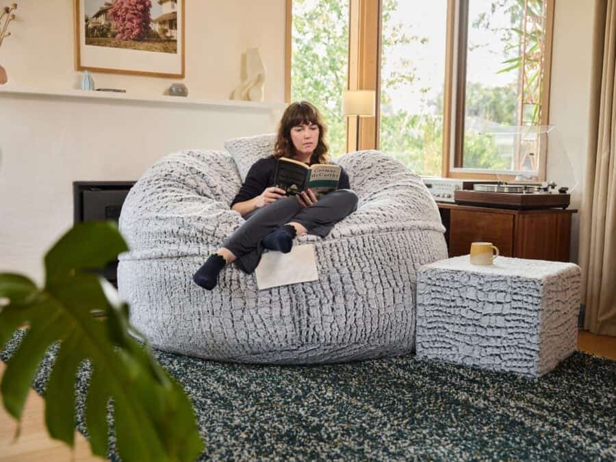 A woman relaxing on a large foam filled Stone Fur Grande bean bag in a bright, modern living room with natural light.