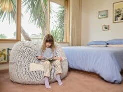 A young girl sitting on a foam filled Stone Fur Kids bean bag reading a book in a bright, airy bedroom.