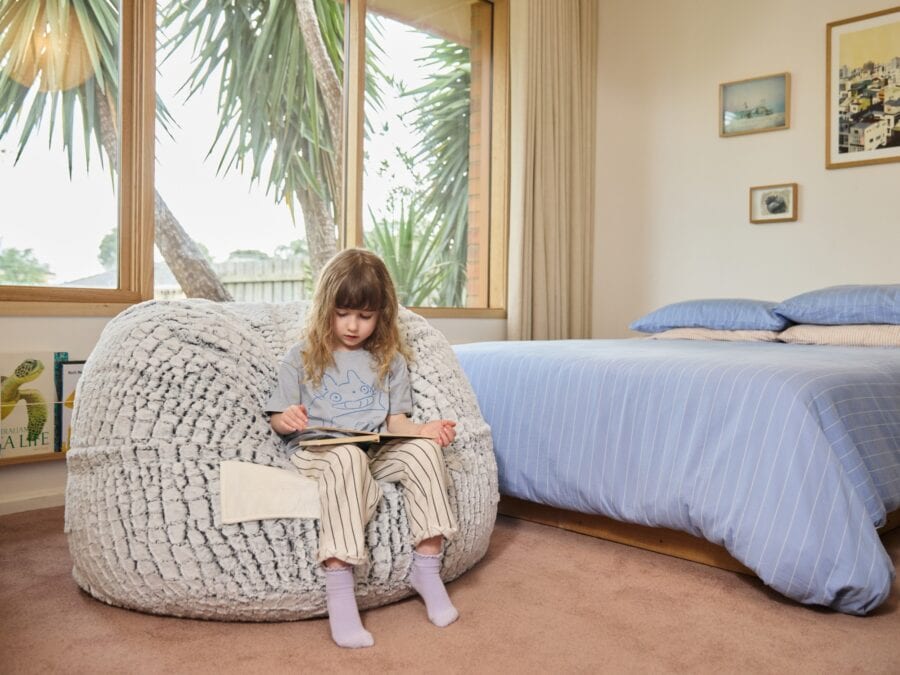 A young girl sitting on a foam filled Stone Fur Kids bean bag reading a book in a bright, airy bedroom.
