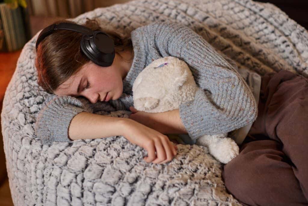 Woman resting on an Urban foam filled bean bag with headphones and teddy bear.