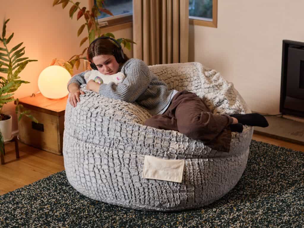 Relaxing teenage girl on a Stone Faux Fur foam filled Urban bean bag in a calm living room setting.